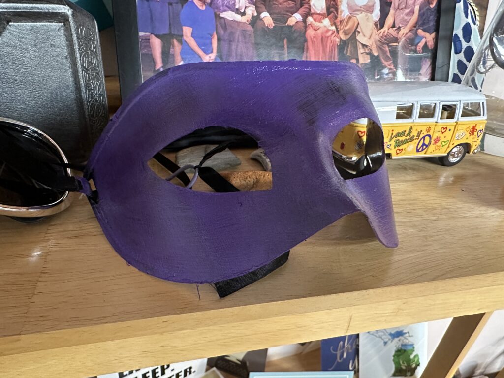 A domino mask, painted purple, sits on a shelf of mementos, including a die-cast VW minibus, a framed photograph, a pair of sunglasses, and a plastic Mjolnir replica.