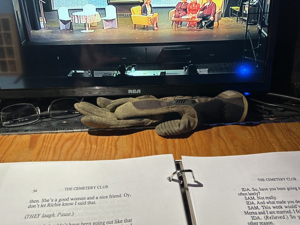An open script for The Cemetery Club sits on a wooden desk below a pair of Mechanix work gloves, eyeglasses, and an RCA video monitor showing a view of the stage. Four actors are visible on stage, three seated on a sofa and the fourth in an armchair.
